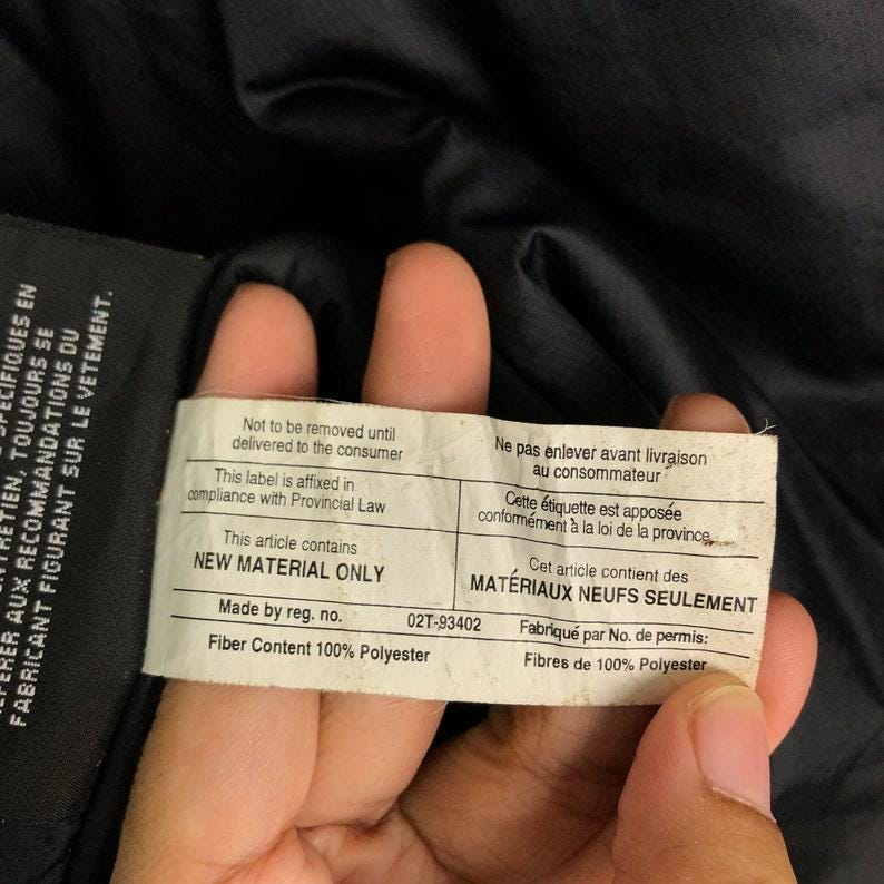 Puede incluir: Primer plano de una etiqueta blanca con texto negro, sostenida sobre un fondo de tela negra. La etiqueta incluye texto en ingl&eacute;s y franc&eacute;s, que indica "NEW MATERIAL ONLY" y "Fiber Content 100% Polyester".
