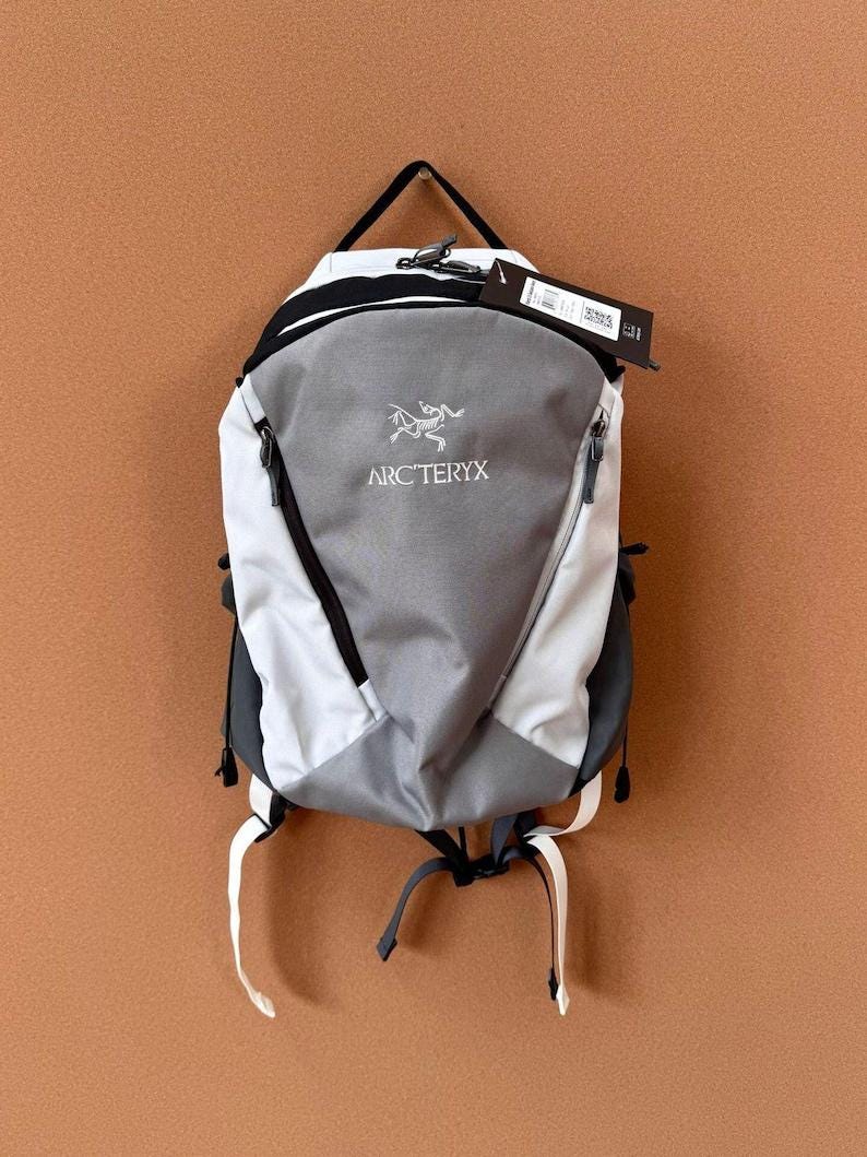 May include: A gray, white, and black backpack with the Arc'teryx logo. The backpack has a top handle, two side pockets, and adjustable straps. The backpack is hanging against a brown background.