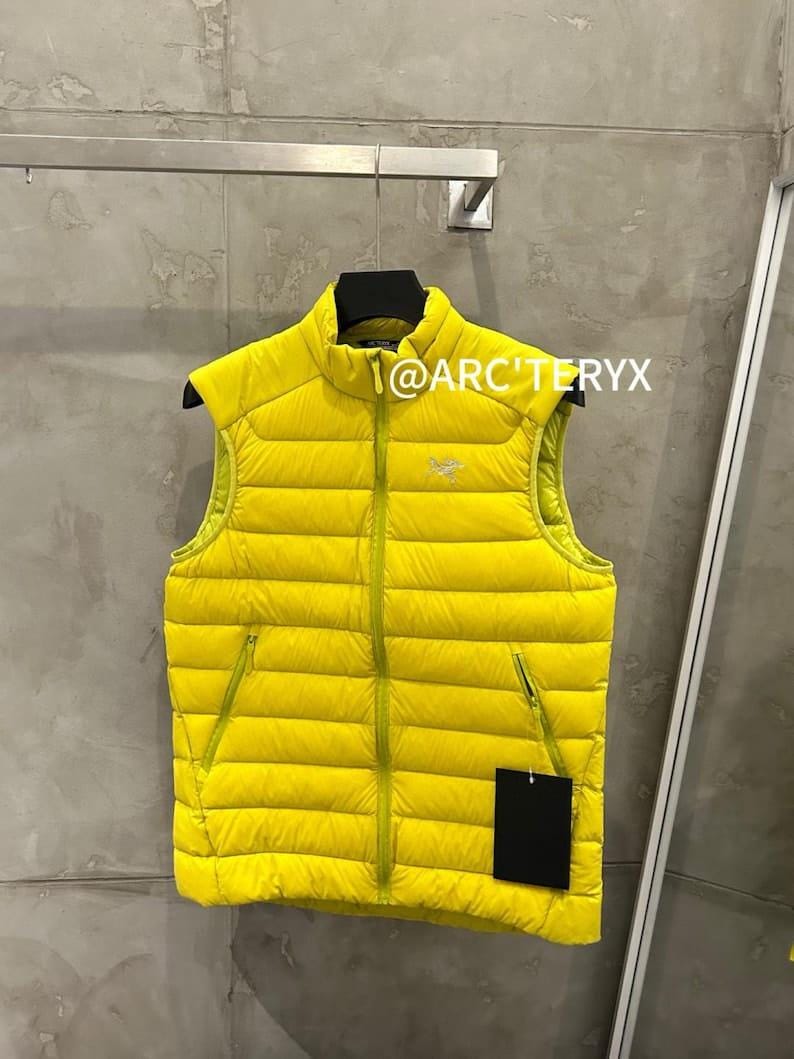 May include: A bright yellow puffer vest with a high collar and a full-length zipper. The vest has two side pockets and a small logo on the chest. The text "@ARC'TERYX" is visible on the top of the vest.