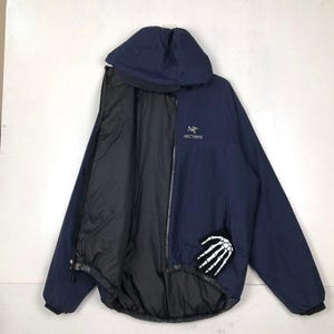 Puede incluir: Chaqueta con capucha azul marino con interior negro. La chaqueta tiene el nombre de la marca "Arc'teryx" en blanco en el pecho y un parche de mano de esqueleto blanco en la parte inferior derecha. La chaqueta est&aacute; abierta, mostrando el forro negro.