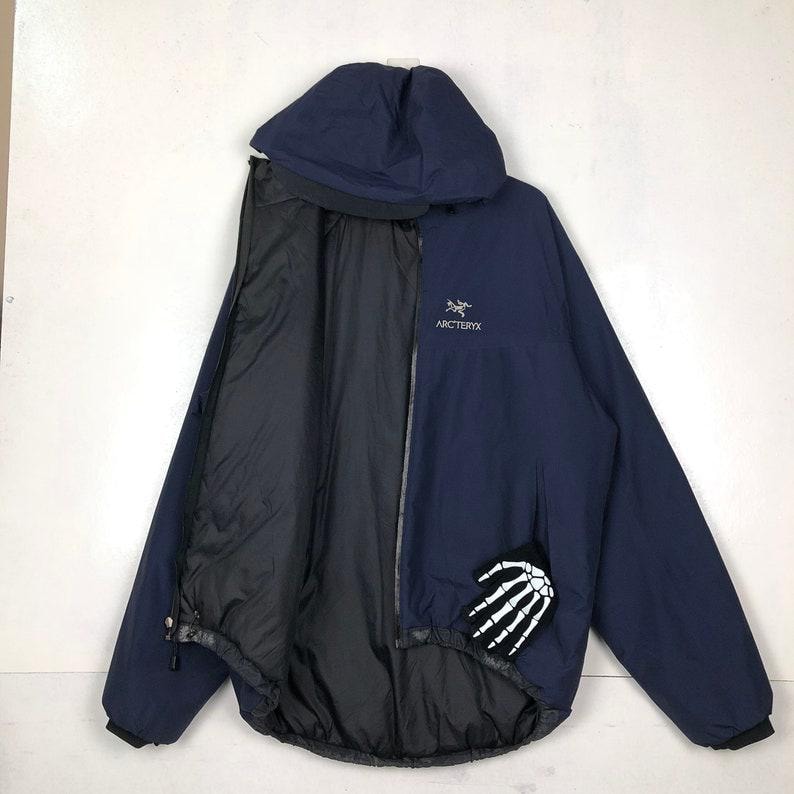 Puede incluir: Chaqueta con capucha azul marino con interior negro. La chaqueta tiene el nombre de la marca "Arc'teryx" en blanco en el pecho y un parche de mano de esqueleto blanco en la parte inferior derecha. La chaqueta est&aacute; abierta, mostrando el forro negro.