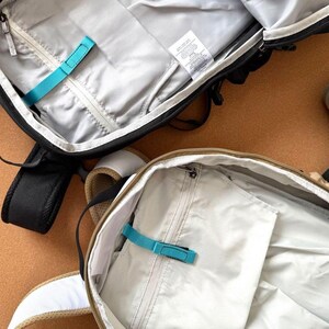 May include: Two open backpacks with interior compartments visible. One is black with a gray interior, a teal strap, and a zipper. The other is tan with a white strap and a gray interior. Both have a tag.