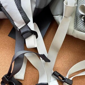May include: Close-up of a backpack with gray and white straps and black plastic buckles. The straps are made of a durable, woven material. The backpack appears to be new and unused.