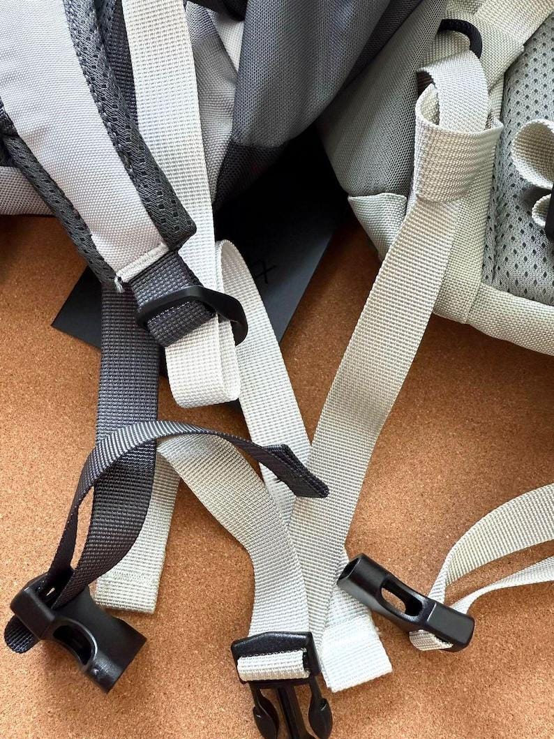 May include: Close-up of a backpack with gray and white straps and black plastic buckles. The straps are made of a durable, woven material. The backpack appears to be new and unused.