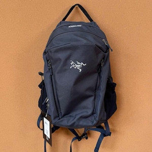 May include: A navy blue backpack with the brand name "Arc'teryx" printed in white. The backpack has a main compartment, side pockets, and adjustable straps. A tag hangs from the straps. The backpack is hanging against a brown wall.