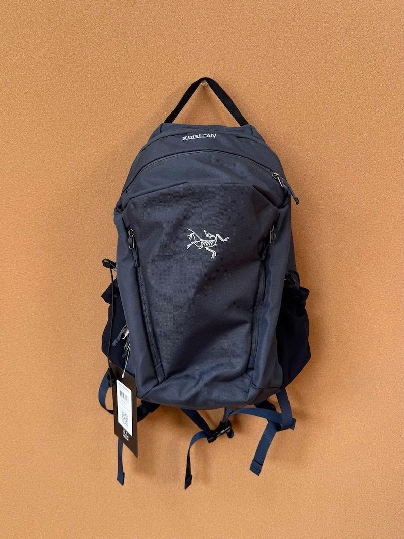 May include: A navy blue backpack with the brand name "Arc'teryx" printed in white. The backpack has a main compartment, side pockets, and adjustable straps. A tag hangs from the straps. The backpack is hanging against a brown wall.