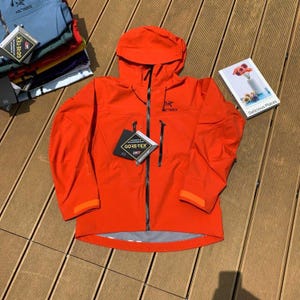 May include: Bright orange hooded jacket with a black zipper and two front pockets. The jacket has the brand name "Arc'teryx" printed on the chest and a "GORE-TEX" tag. Additional jackets in various colors are visible in the background.