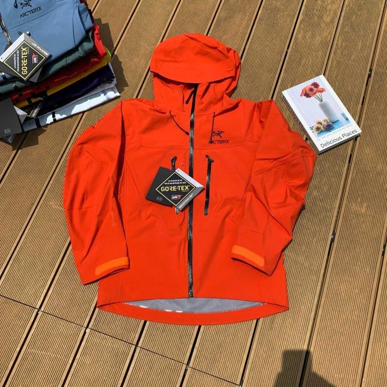May include: Bright orange hooded jacket with a black zipper and two front pockets. The jacket has the brand name "Arc'teryx" printed on the chest and a "GORE-TEX" tag. Additional jackets in various colors are visible in the background.