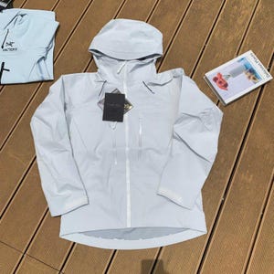 May include: A light blue hooded jacket with a full-length zipper and two front pockets. The jacket has a tag attached and is laid out on a wooden surface. A folded light blue shirt is visible in the background.