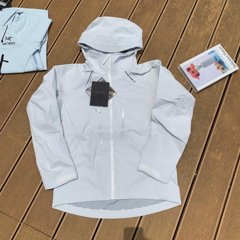 May include: A light blue hooded jacket with a full-length zipper and two front pockets. The jacket has a tag attached and is laid out on a wooden surface. A folded light blue shirt is visible in the background.