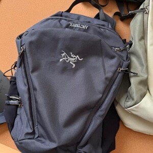 May include: A dark gray backpack with the brand name "ARCTERYX" and a white logo. The backpack has a top handle, two zippered compartments, and adjustable straps. Another backpack is partially visible in the background.