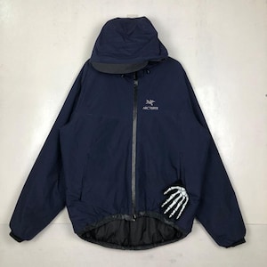 Puede incluir: Chaqueta con capucha azul marino con cremallera completa y el nombre de la marca "Arc'teryx" bordado en el pecho. La chaqueta presenta un forro negro y un parche de mano de esqueleto blanco en la manga.