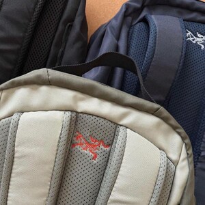 May include: Close-up of three backpacks in various colors. The front-most backpack is tan with a red logo. The other two backpacks are black and navy blue. All three backpacks have padded backs and shoulder straps.