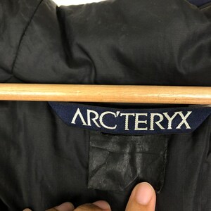 Puede incluir: Primer plano de una chaqueta negra con el nombre de la marca ARC'TERYX en letras blancas sobre una etiqueta azul marino. La chaqueta est&aacute; colgada de una percha de madera.