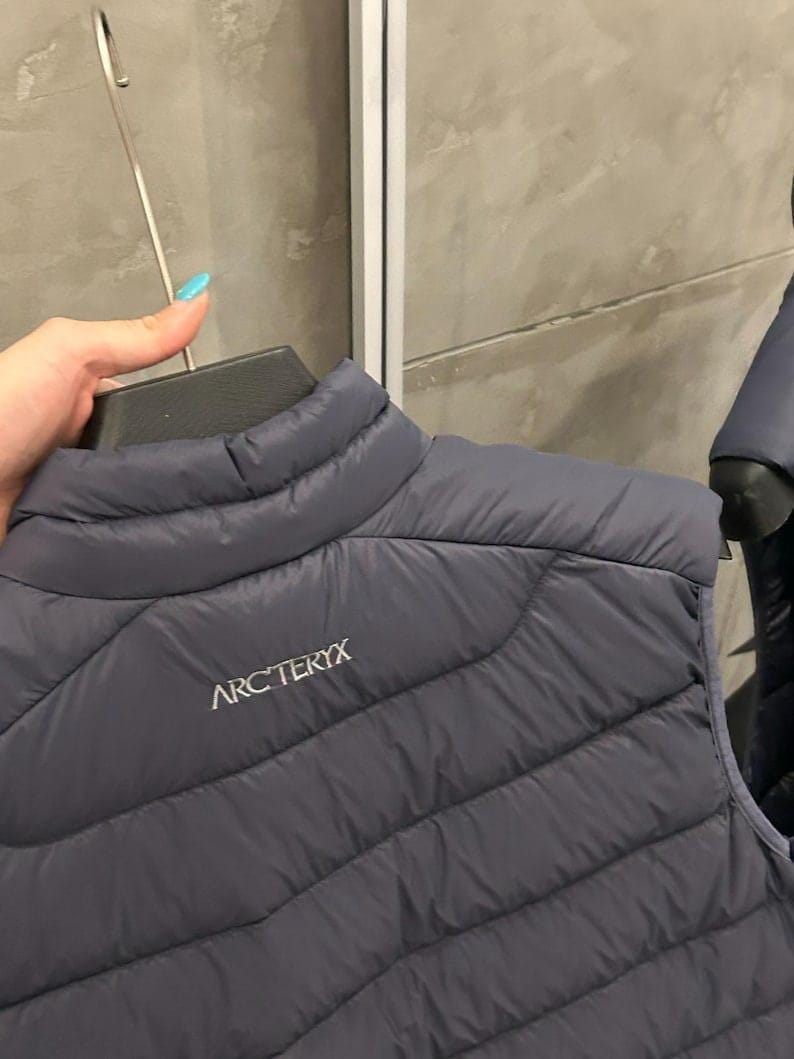 May include: A dark gray quilted vest with the brand name "ARC'TERYX" printed in white. The vest is hanging on a black hanger. The vest has a high collar and a diagonal quilted pattern.
