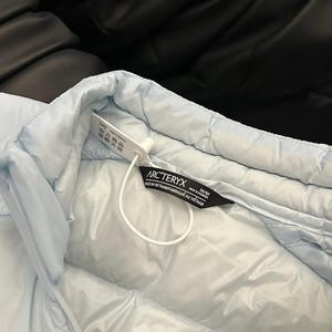 May include: Light blue quilted jacket with a tag that reads "ARC'TERYX". The jacket is made of a smooth, padded material. The tag also indicates the jacket is made in Vietnam.