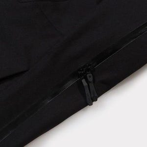 May include: Close-up of a black garment with a zipper. The zipper is black and runs diagonally across the image. The fabric appears to be a smooth, matte material. The background is white.