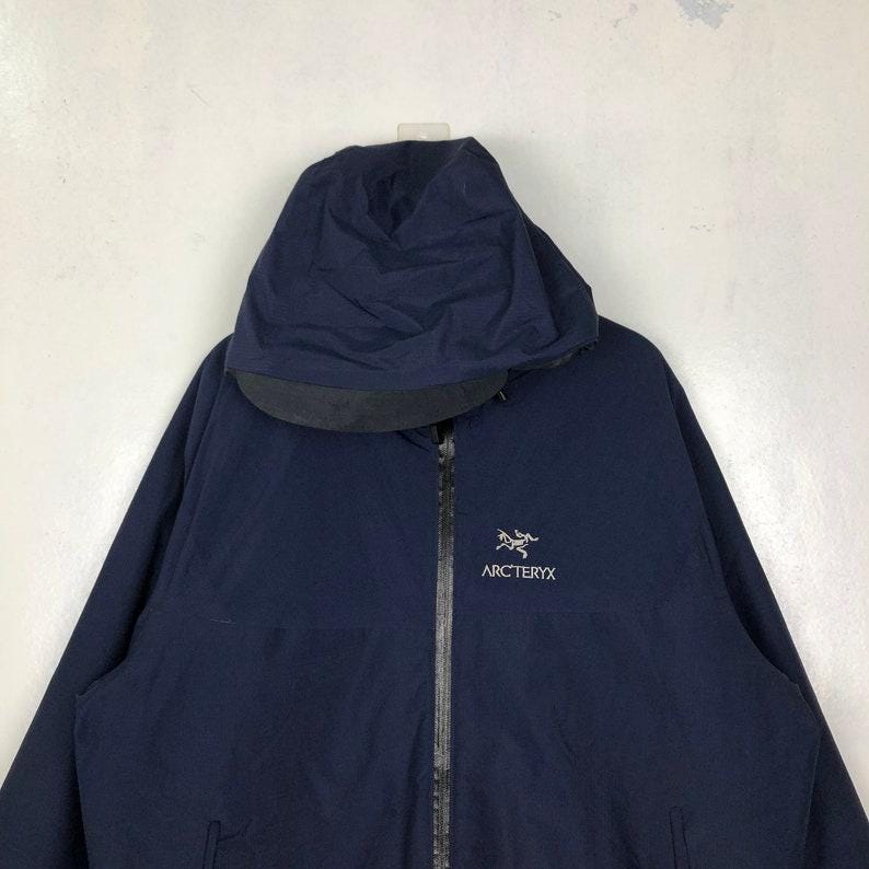 Puede incluir: Chaqueta azul marino con capucha y cremallera completa. La marca "Arc'teryx" est&aacute; en blanco en el pecho. La capucha est&aacute; doblada, revelando un forro negro. La chaqueta parece estar hecha de un material liso, posiblemente resistente al agua.