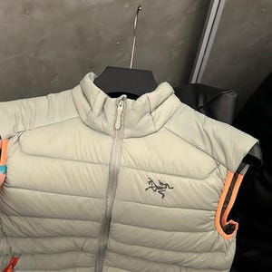 May include: A light gray puffer vest with a full-length zipper and orange trim. The vest has a small logo on the chest and is hanging on a black hanger. The vest is made of a quilted material.