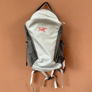May include: A gray and light green backpack with white straps and a black handle. The backpack features a red logo and multiple zippers. The bag is designed for outdoor activities and travel.