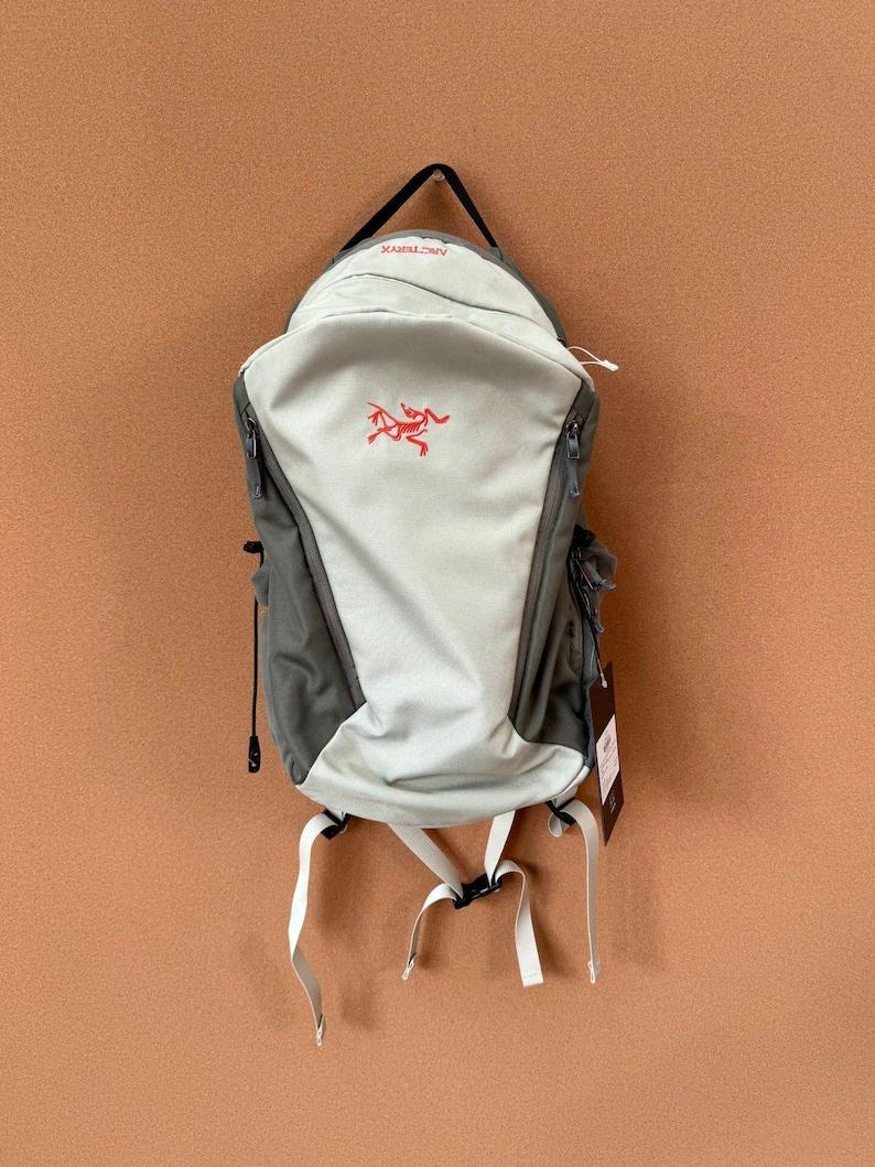 May include: A gray and light green backpack with white straps and a black handle. The backpack features a red logo and multiple zippers. The bag is designed for outdoor activities and travel.