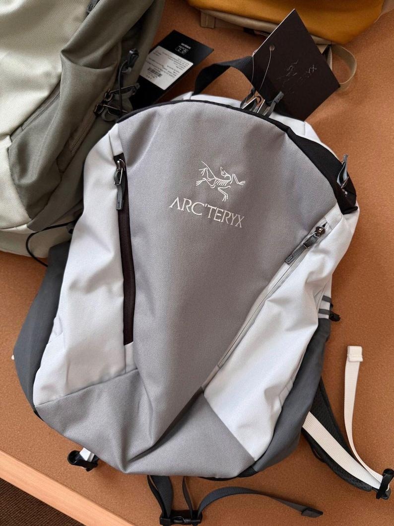 May include: A gray and white backpack with the Arc'teryx logo. The backpack has a zippered main compartment and a smaller front pocket. The straps are black and white. The backpack is made of a durable, water-resistant material.