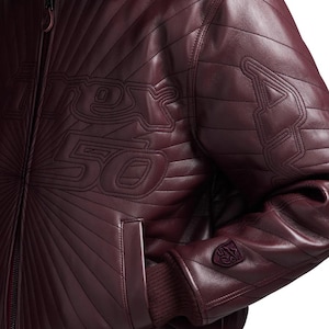 K&ouml;nnte beinhalten: Burgunderfarbene Lederjacke mit Steppdetails und dem Text "Rex 50" auf der Brust. Sie hat gerippte B&uuml;ndchen und ein kleines Emblem am &Auml;rmel. Die Jacke hat einen Rei&szlig;verschluss und einen klassischen Bomberstil.