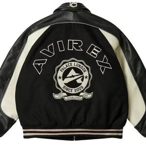 Op de afbeelding: Zwarte en cr&egrave;mekleurige varsity jas met leren mouwen en een zwarte body. De achterkant toont het woord "AVIREX" in witte letters boven een cirkelvormig embleem met "PALACE LONDON SINCE 2009 ESTABLISHED 1975".