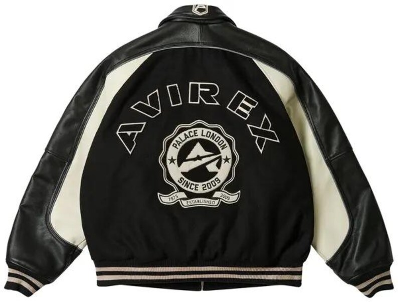 Op de afbeelding: Zwarte en cr&egrave;mekleurige varsity jas met leren mouwen en een zwarte body. De achterkant toont het woord "AVIREX" in witte letters boven een cirkelvormig embleem met "PALACE LONDON SINCE 2009 ESTABLISHED 1975".