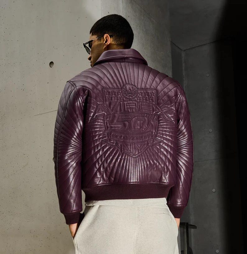 K&ouml;nnte beinhalten: Burgunderfarbene Lederjacke mit Steppmuster, geripptem Kragen und B&uuml;ndchen. Auf der R&uuml;ckseite befindet sich ein gepr&auml;gtes Design mit dem Text "VRC 50". Die Jacke wird mit hellgrauer Jogginghose getragen.