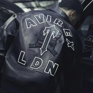 May include: Black leather jacket with white lettering and a logo on the back. The jacket features the words "AVIREX" and "LDN" in a bold, outlined font. A large logo is centered on the back, with the letter "T" prominently displayed.
