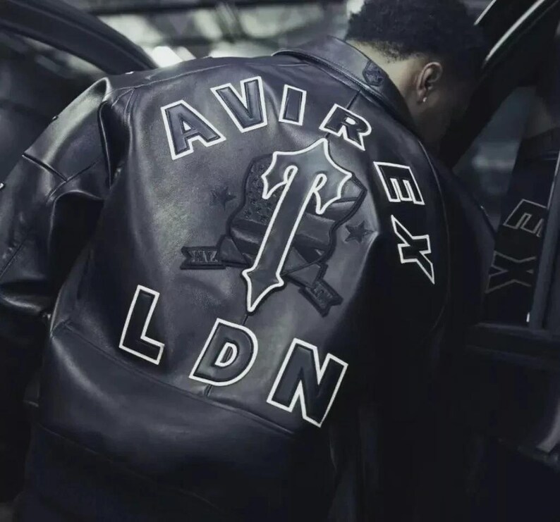 May include: Black leather jacket with white lettering and a logo on the back. The jacket features the words "AVIREX" and "LDN" in a bold, outlined font. A large logo is centered on the back, with the letter "T" prominently displayed.