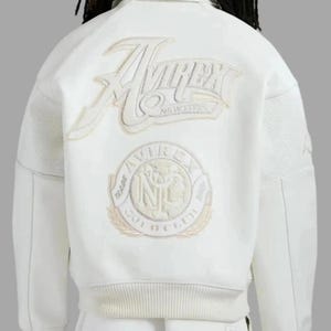May include: Cream leather jacket with embroidered "Avirex New York" logo on the back. Features a circular patch with text and a laurel wreath design. Includes mesh paneling on the sides.