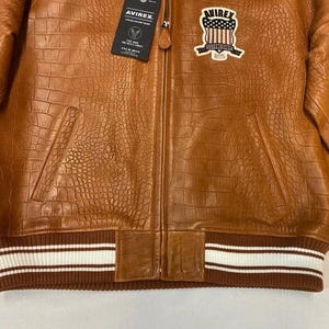 May include: A brown leather jacket with a crocodile texture. The jacket has a zipper closure, two front pockets, and a ribbed hem with white stripes. A patch with a shield design is on the chest.