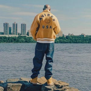 May include: A mustard yellow leather jacket with the word "AVIREX" and "USA" printed on the back. The jacket is paired with dark blue denim jeans and tan work boots. The person is standing on a rock formation near a body of water with a city skyline in the background.