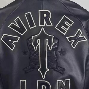 May include: Black leather jacket with white embroidered lettering and patches. The back of the jacket features the word "AVIREX" across the top, a central emblem, and the letters "LDN" at the bottom. The emblem includes a cross-shaped design and a shield.