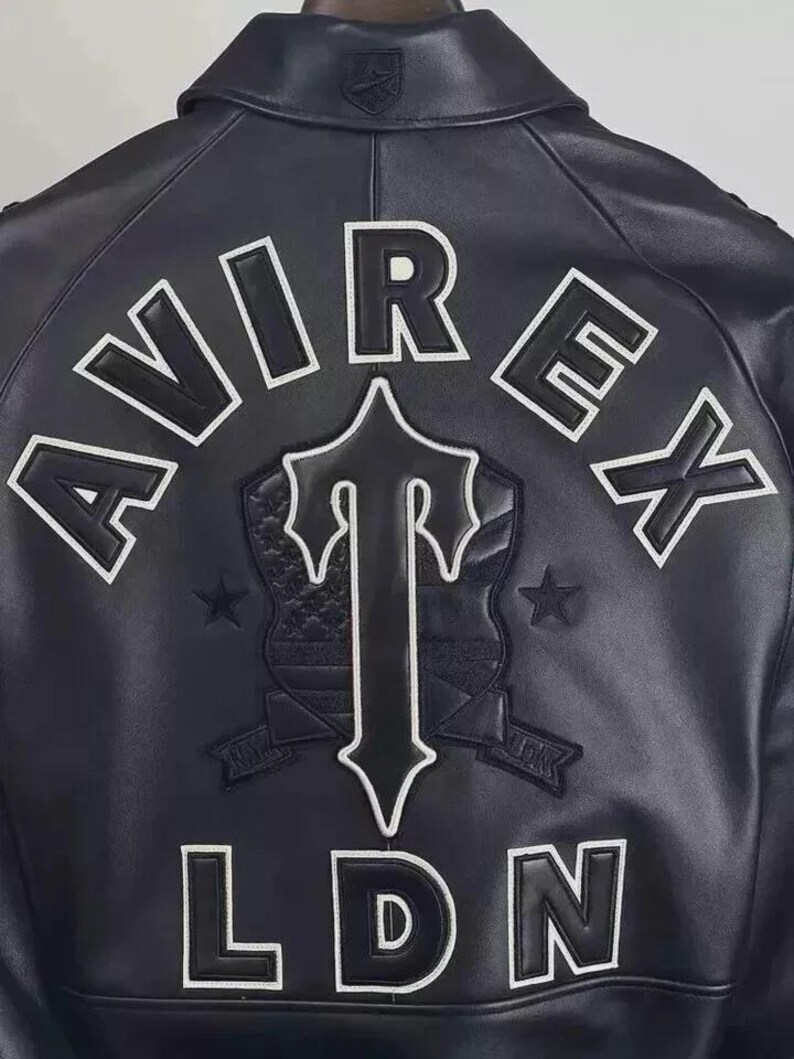 May include: Black leather jacket with white embroidered lettering and patches. The back of the jacket features the word "AVIREX" across the top, a central emblem, and the letters "LDN" at the bottom. The emblem includes a cross-shaped design and a shield.