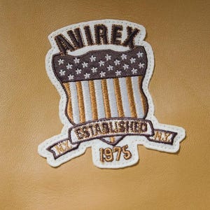 May include: Embroidered patch with the text "AVIREX" at the top, a shield design with stars and stripes, and the words "ESTABLISHED NY 1975" on a banner. The patch is white, brown, gold, and blue.