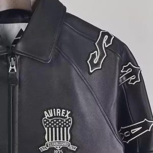 May include: Black leather jacket with white embroidered patches and the "AVIREX" logo. The jacket features a zipper closure and a collared neckline. The patches include stylized lettering and a shield design with stars and stripes. "ESTABLISHED NY 1975" is also visible.