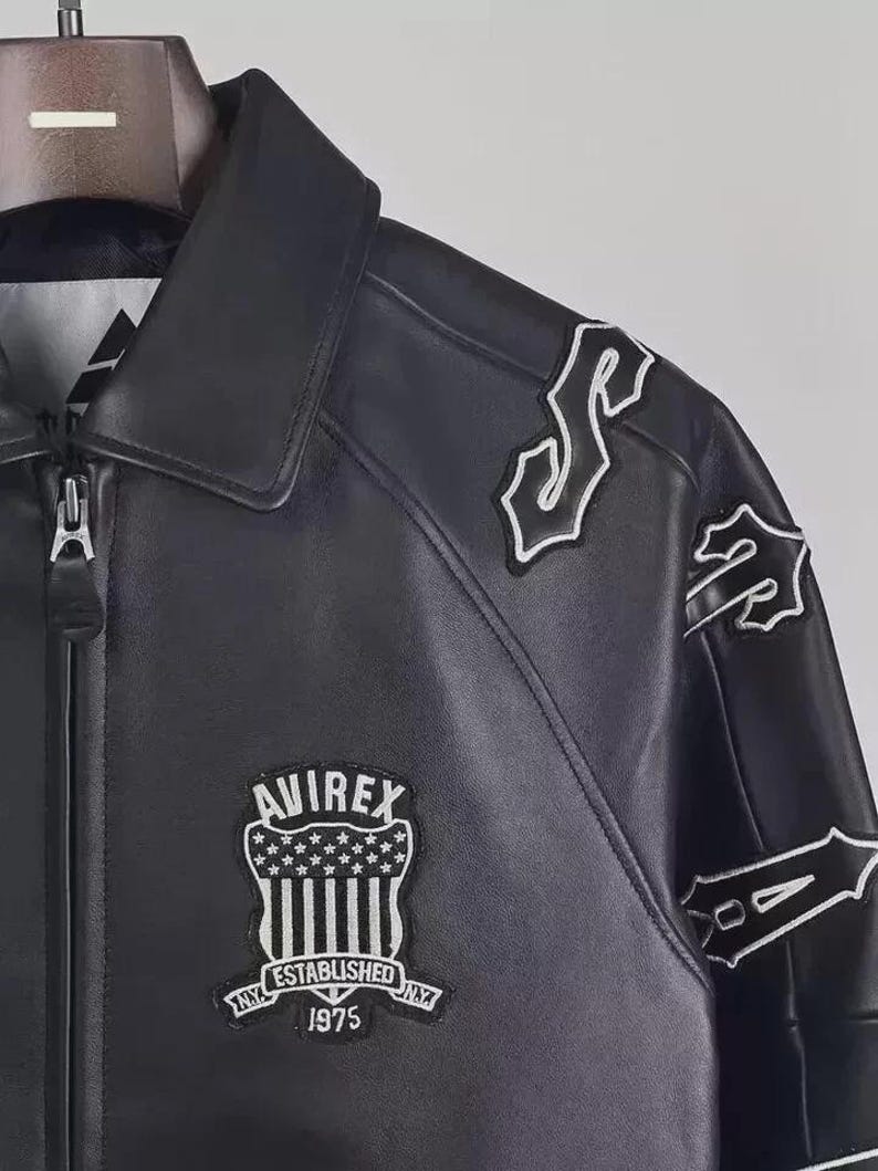 May include: Black leather jacket with white embroidered patches and the "AVIREX" logo. The jacket features a zipper closure and a collared neckline. The patches include stylized lettering and a shield design with stars and stripes. "ESTABLISHED NY 1975" is also visible.