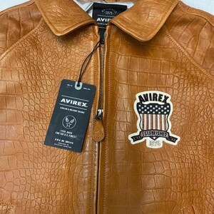 May include: A brown leather jacket with a textured pattern, a zipper closure, and a collar. The jacket features a patch with the text "AVIREX ESTABLISHED 1975" and a tag with the brand logo and text. The jacket is a Type AVRX for the U.S. Finest.