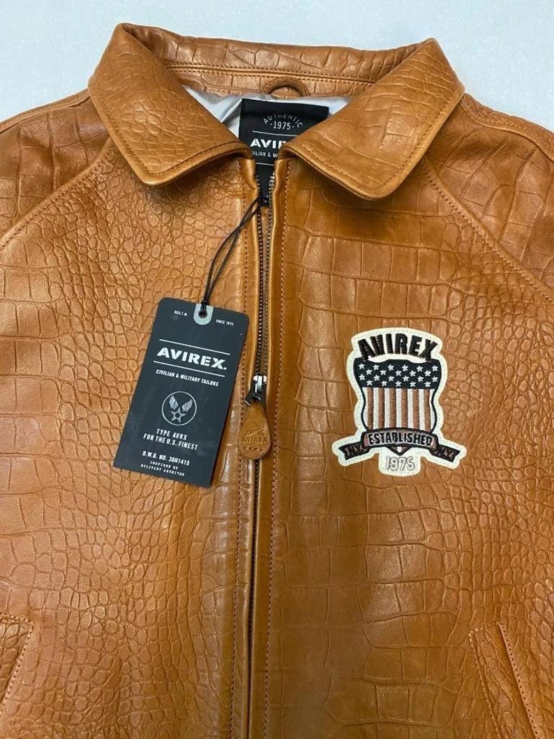 May include: A brown leather jacket with a textured pattern, a zipper closure, and a collar. The jacket features a patch with the text "AVIREX ESTABLISHED 1975" and a tag with the brand logo and text. The jacket is a Type AVRX for the U.S. Finest.
