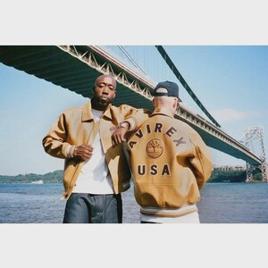 May include: Two people wearing tan leather jackets with the text "AVIREX USA" on the back. The jackets have a ribbed hem and cuffs. The background includes a bridge and water.