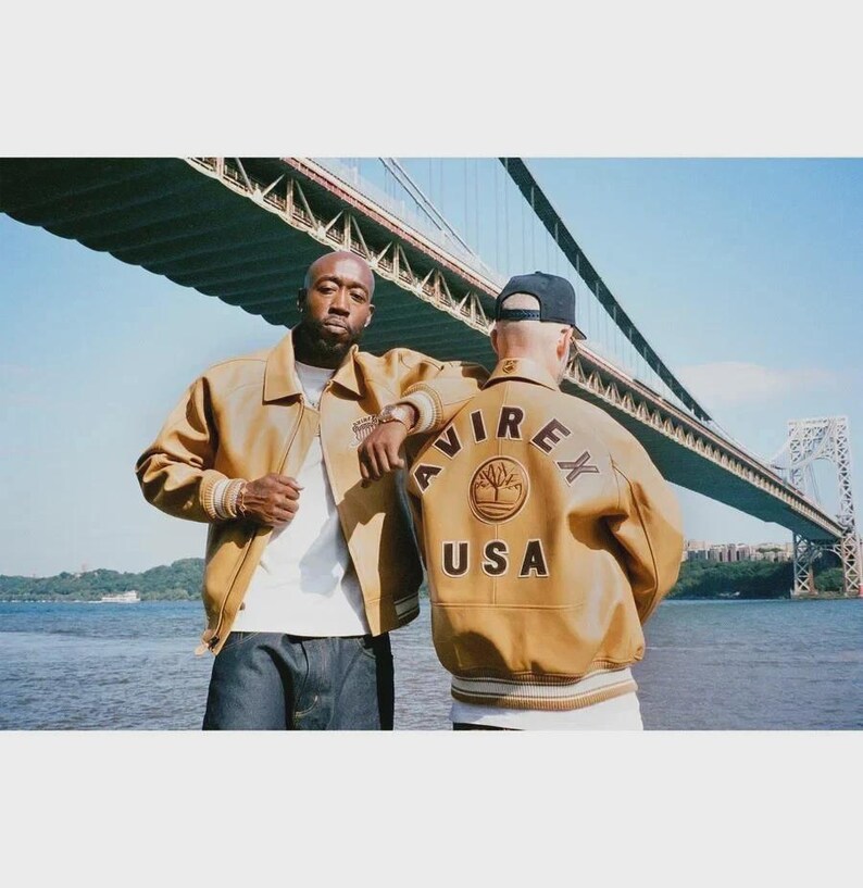 May include: Two people wearing tan leather jackets with the text "AVIREX USA" on the back. The jackets have a ribbed hem and cuffs. The background includes a bridge and water.