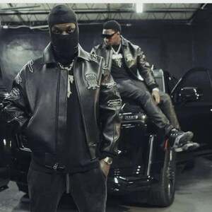 May include: Two people wearing black leather jackets and black clothing. One person is wearing a black balaclava. The jackets have patches on the sleeves and chest. They are standing and sitting near a black vehicle.