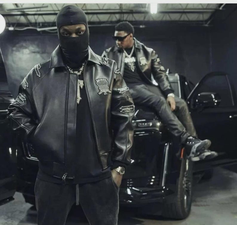 May include: Two people wearing black leather jackets and black clothing. One person is wearing a black balaclava. The jackets have patches on the sleeves and chest. They are standing and sitting near a black vehicle.