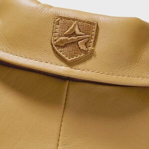 May include: Close-up of a tan leather item with a stitched emblem. The emblem is a shield shape with a detailed embroidered design in a matching tan color. The leather has a smooth texture.