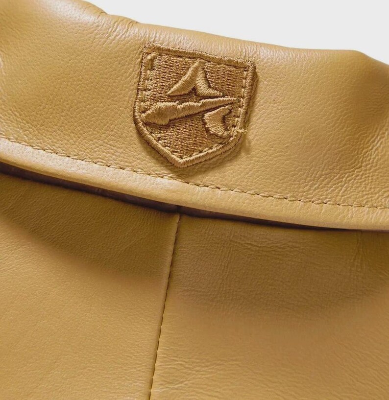 May include: Close-up of a tan leather item with a stitched emblem. The emblem is a shield shape with a detailed embroidered design in a matching tan color. The leather has a smooth texture.