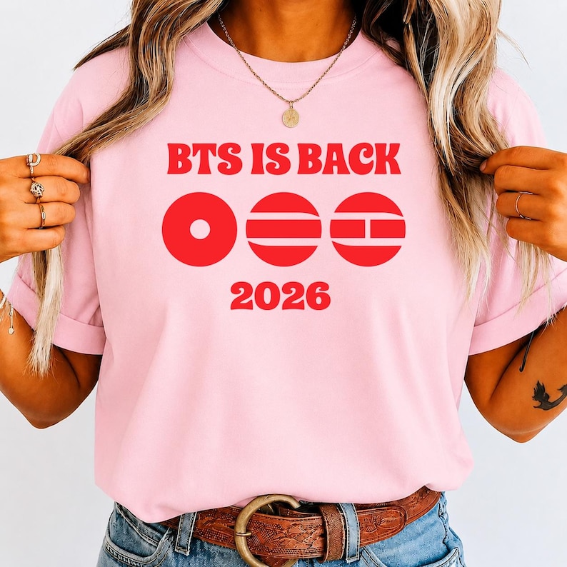 May include: A light pink t-shirt featuring the red text "BTS IS BACK" at the top, followed by three red circular graphic icons with horizontal and vertical lines, and the red text "2026" at the bottom, all printed on the front of the garment.