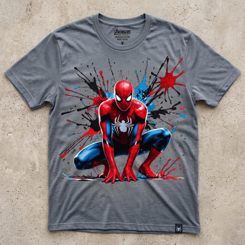 May include: A heather gray short-sleeve t-shirt featuring a graphic of Spider-Man in a crouching pose. The design includes red, blue, and black paint splatter effects. The inner neck label reads Avengers Collective, Premium Cotton, Made in USA.